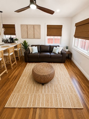 A modern living room with a brown leather sofa, a round woven ottoman, a dining area with wooden chairs, a kitchen with a white countertop, and a ceiling fan. - Sleeps 8 • King Suite • 2 Queens (Chula Vista)