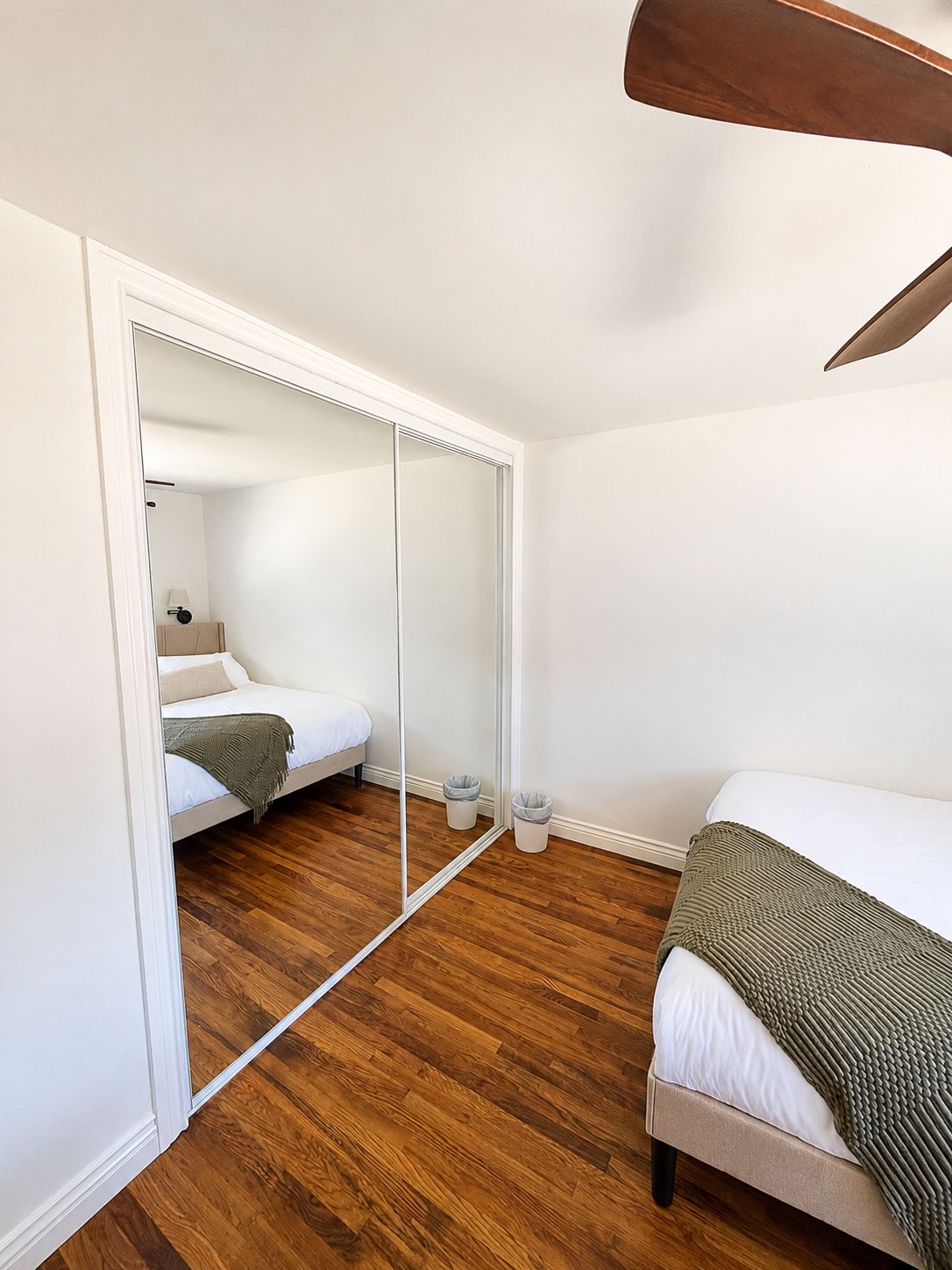 A bedroom with a large mirror, wooden flooring, two beds, and a ceiling fan.