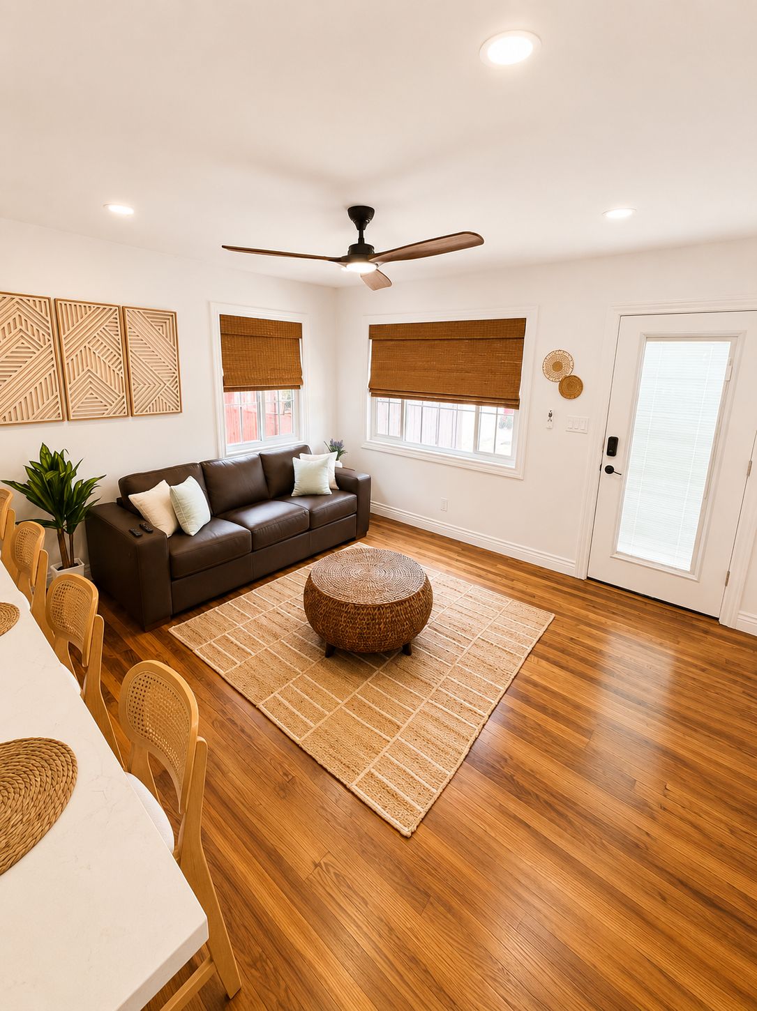 A living room with a brown leather sofa, a round woven ottoman, a dining table with chairs, and a ceiling fan.