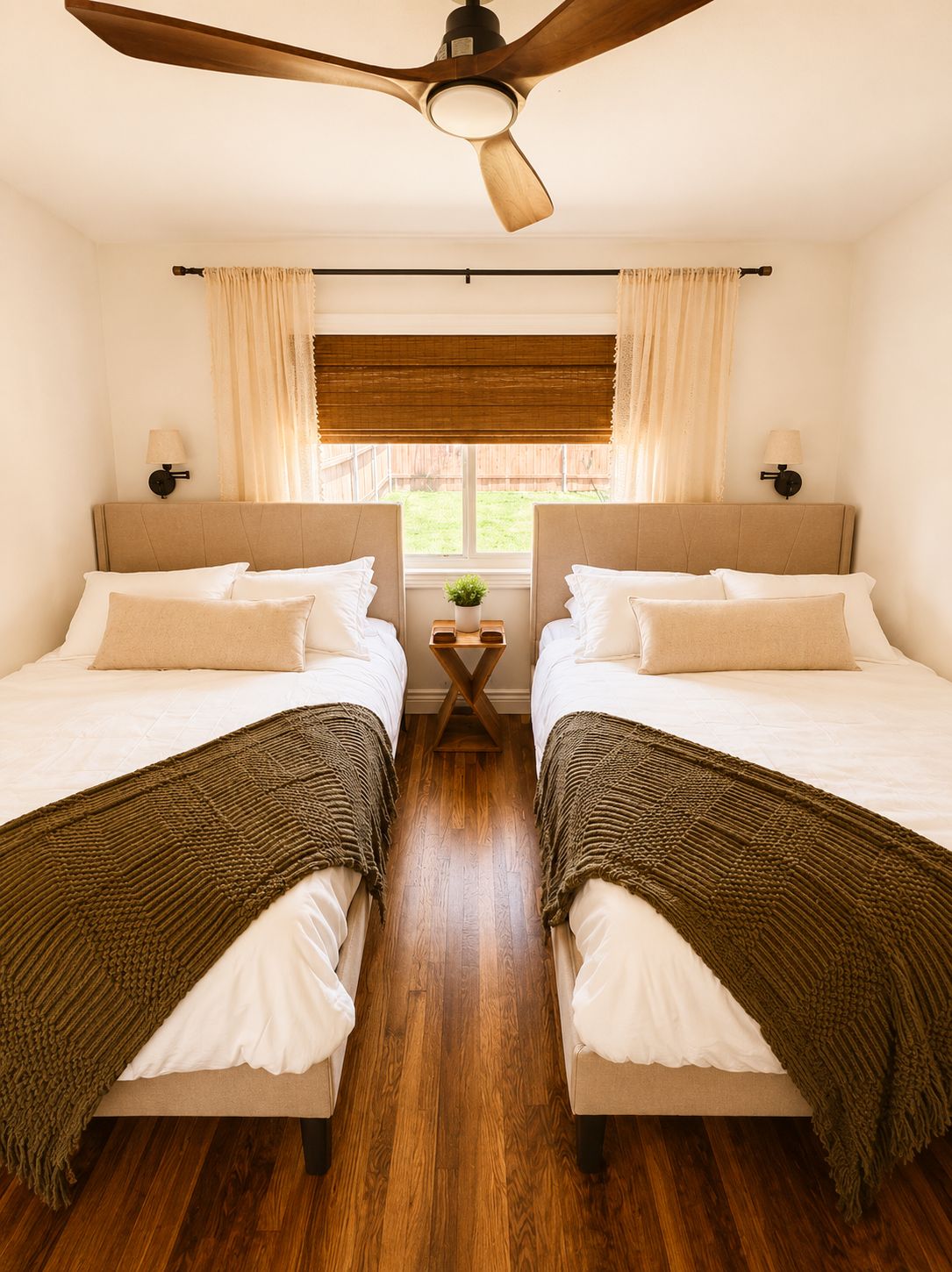 A double room with two beds, a ceiling fan, a small table with a plant, and a window with curtains.