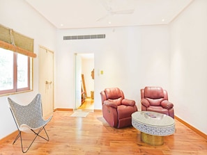 A room with a wooden floor, a patterned chair, two leather armchairs, and a round table with a patterned top. - Suite With Garden View Parijat Villas (Udaipur)