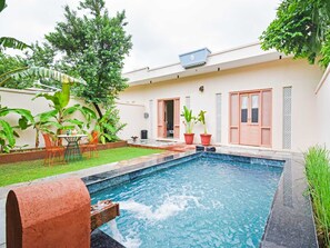 A swimming pool with a water feature, surrounded by a garden with potted plants and a seating area with chairs.