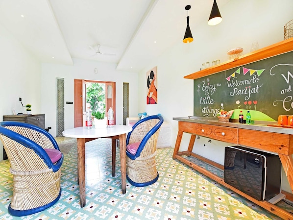 A room with a dining table, two wicker chairs, a small refrigerator, and a chalkboard with a welcome message.
