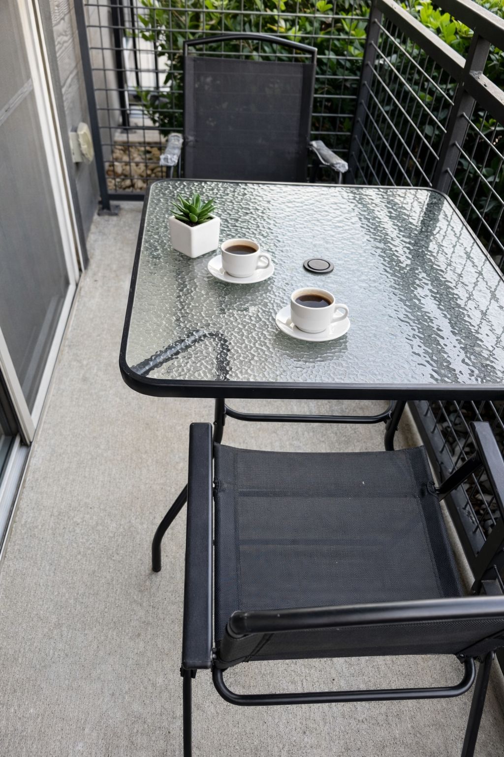 A glass-top table with two white cups of coffee and a small potted plant on a balcony.