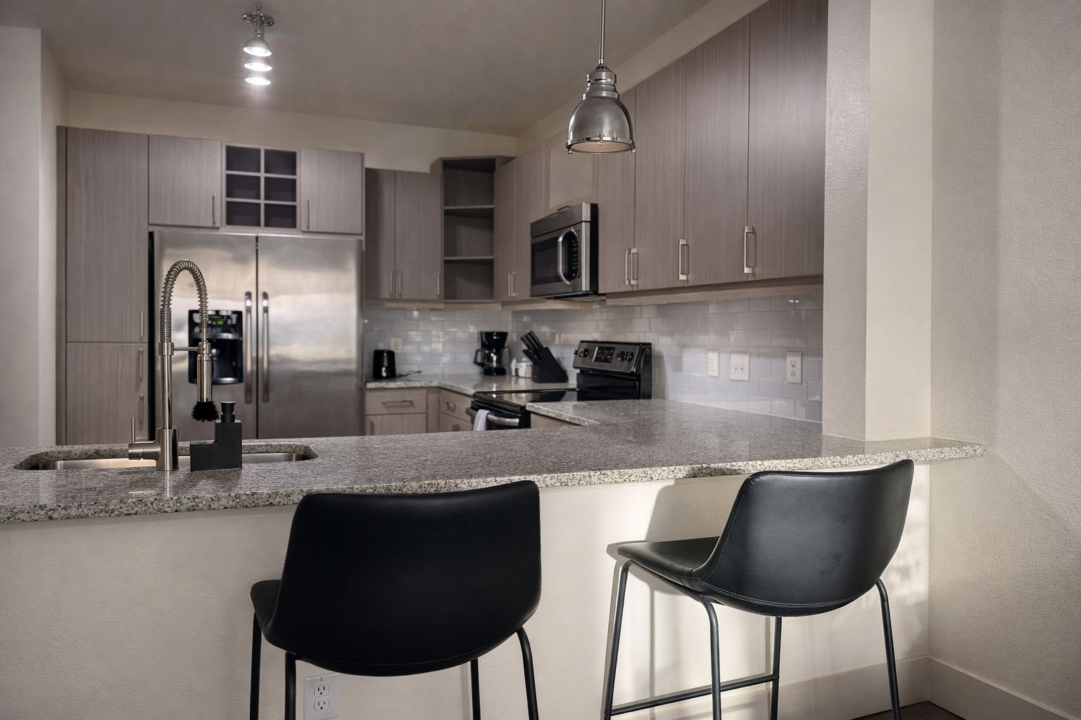 A modern kitchen with stainless steel appliances, a granite countertop, and two black bar stools.