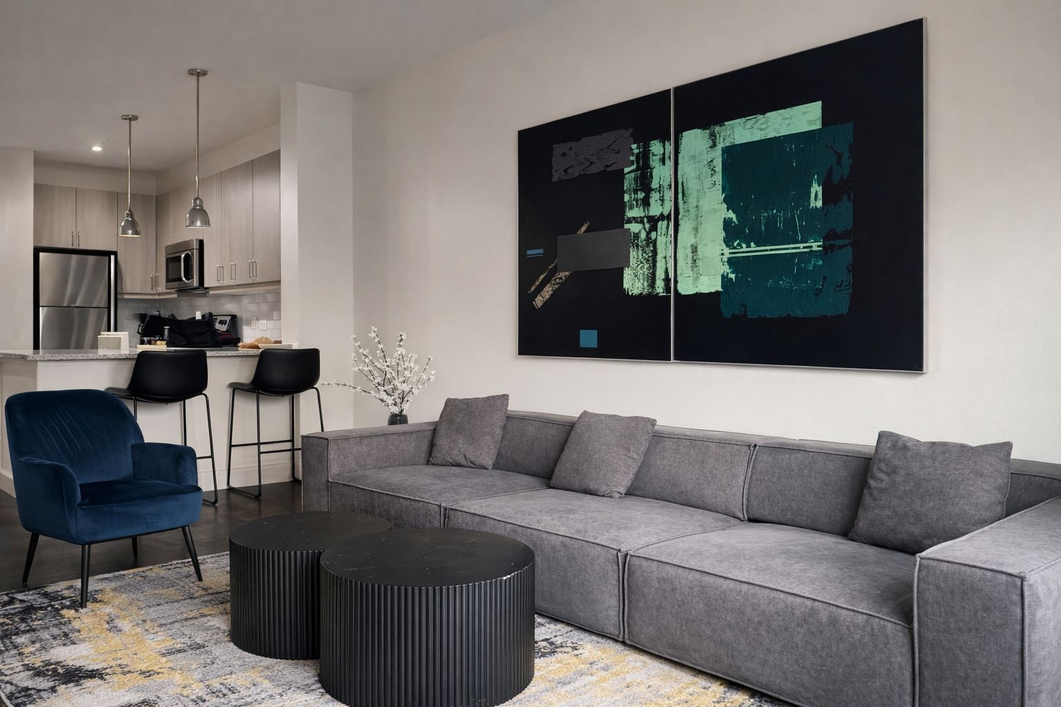 A modern living room with a grey sofa, a blue armchair, and a black coffee table. There is a kitchen in the background with stainless steel appliances.