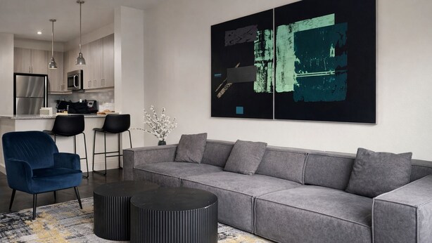 Una sala de estar moderna con un sofá gris, un sillón azul y una mesa de centro negra. Se observa una cocina al fondo con electrodomésticos de acero inoxidable.