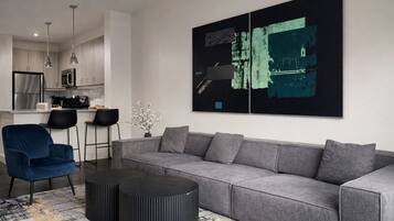 A modern living room with a grey sofa, a blue armchair, and a black coffee table. There is a kitchen in the background with stainless steel appliances.
