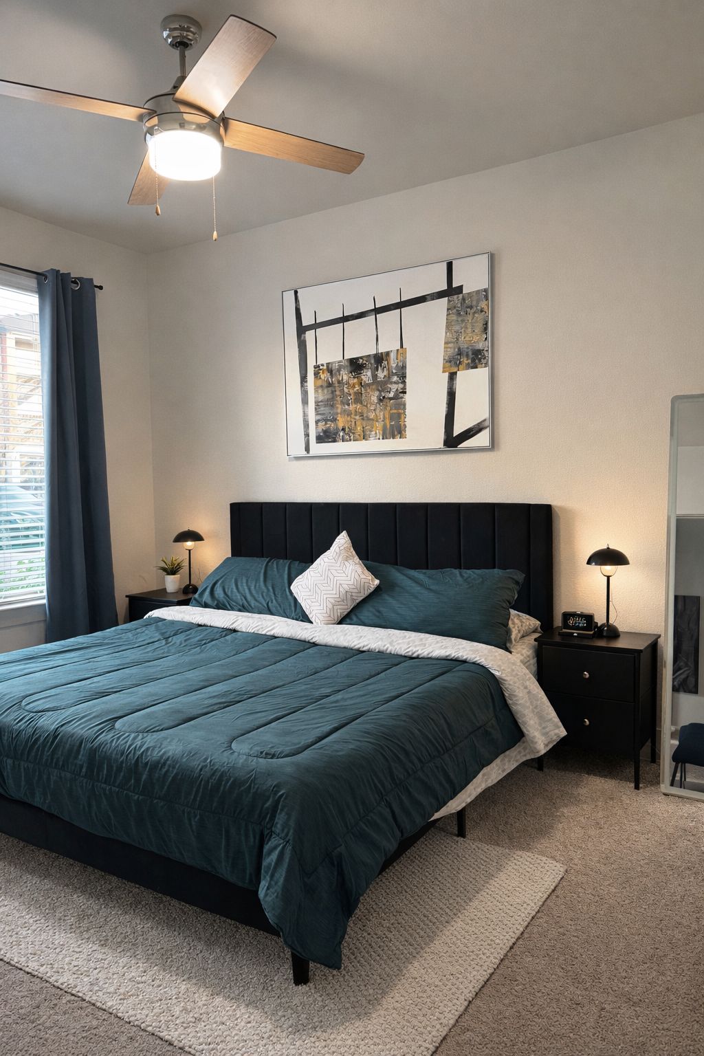 A bedroom with a bed, a ceiling fan, a wall-mounted abstract art piece, and a nightstand with a lamp.