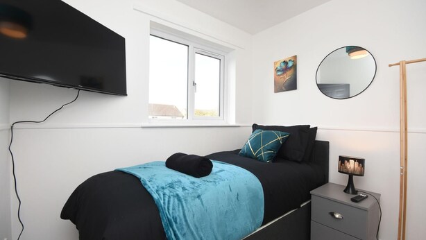 A bedroom with a bed, a bedside table, a lamp, a TV mounted on the wall, and a window. - Beautiful 3BRHome with a large garden in Colchester (Colchester)