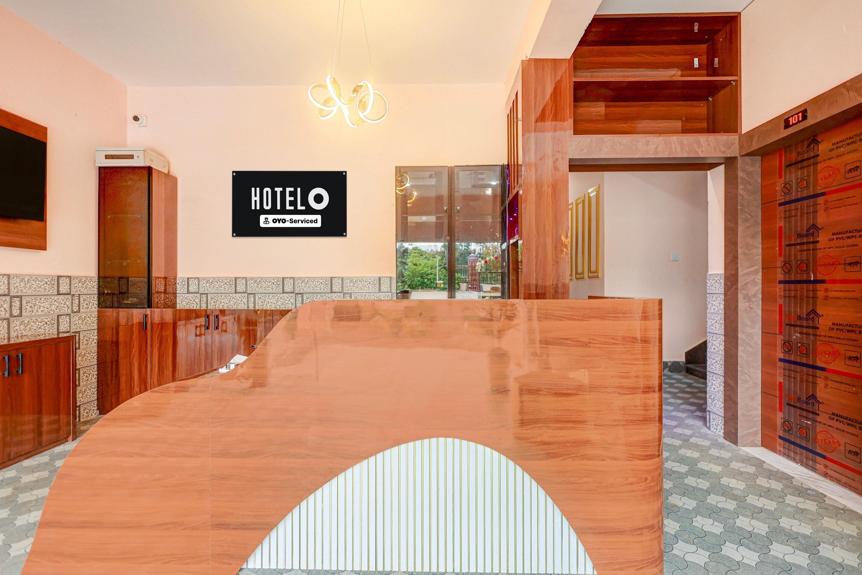 A hotel reception area with a wooden reception desk, a sign indicating it is oyo-serviced, and a view of the outdoors through a glass door.