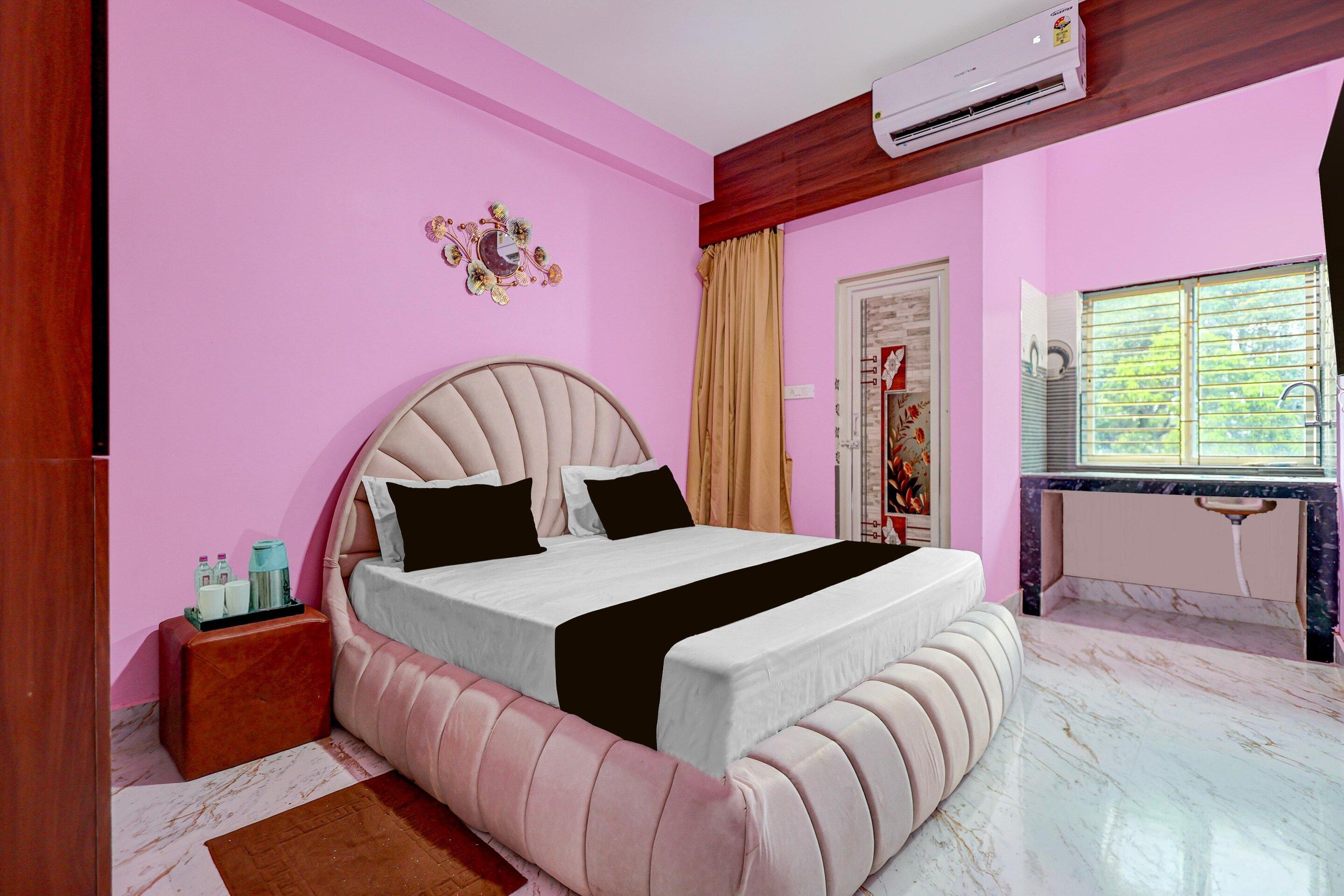 A bedroom with a bed, a bedside table, an air conditioning unit, a window with blinds, and a decorative mirror on the wall.