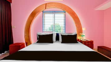 Ein Hotelzimmer mit einem großen Bett, einem Fenster mit horizontalen Rollläden, einem Nachttisch mit Lampe und einem kleinen roten Stuhl.