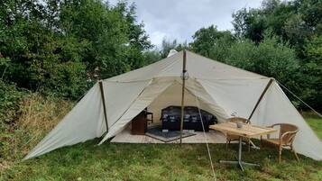 Una carpa con una cama y una mesa, rodeada de pasto y árboles.