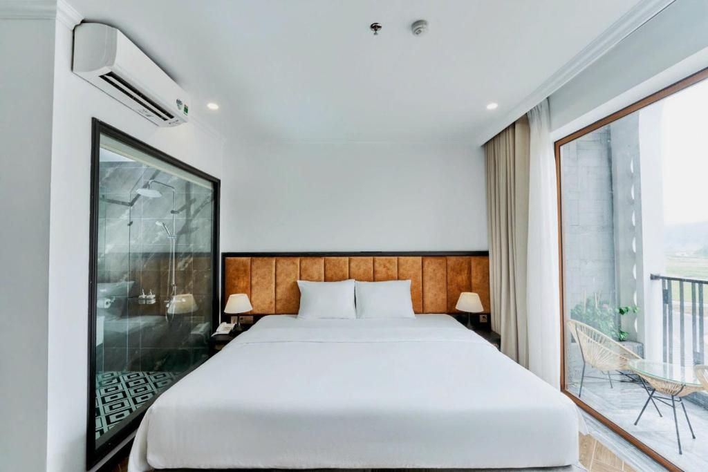 A hotel room with a large bed, an air conditioning unit, a glass shower enclosure, and a balcony with outdoor furniture.