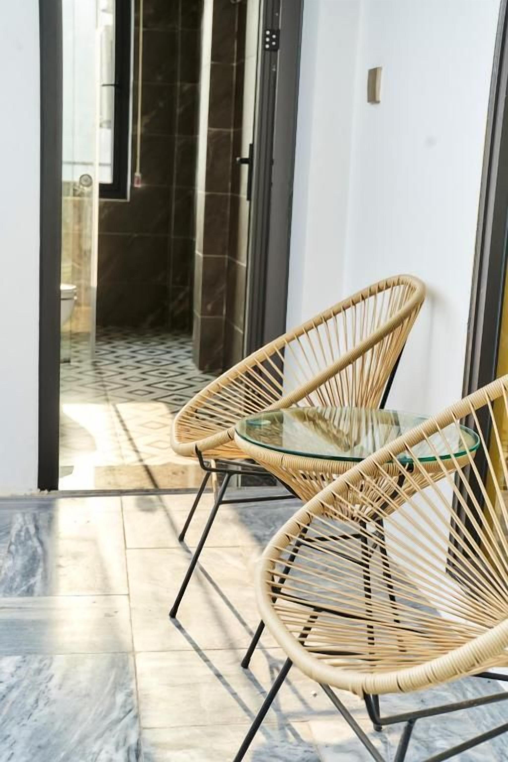 A room with two wicker chairs and a glass table, a tiled floor, and a view of a bathroom with a glass door.