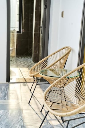 Una habitación con dos sillones de mimbre, una mesa de cristal, suelo de baldosas y vistas a un baño con puerta de cristal.