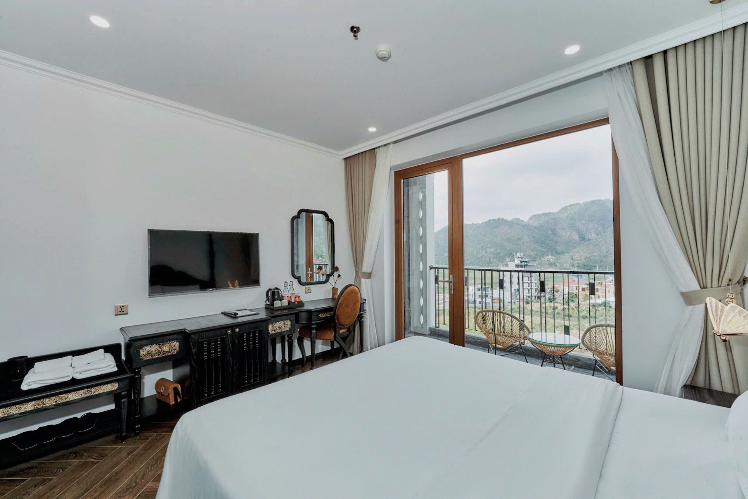 A hotel room with a bed, a desk with a chair, a television, and a balcony with a view of the mountains.