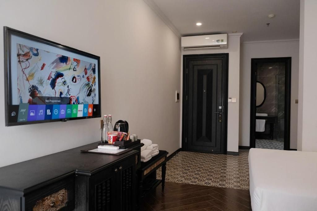 A hotel room with a flat-screen TV, a desk with a coffee maker, a bed, a door leading to a bathroom, and a tiled floor.