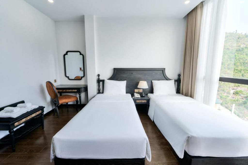 A hotel room with two single beds, a desk with a chair, a mirror, and a view of greenery outside.