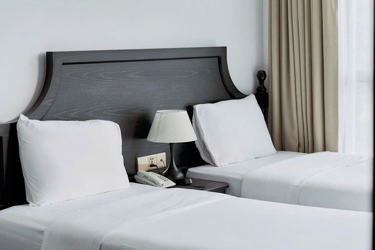 Habitación de hotel con dos camas, una mesita de noche con lámpara y una ventana con cortinas.
