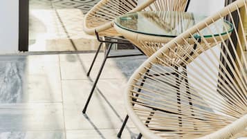 A corridor with a glass door leading to a bathroom, two wicker chairs with a glass table, and a tiled floor.
