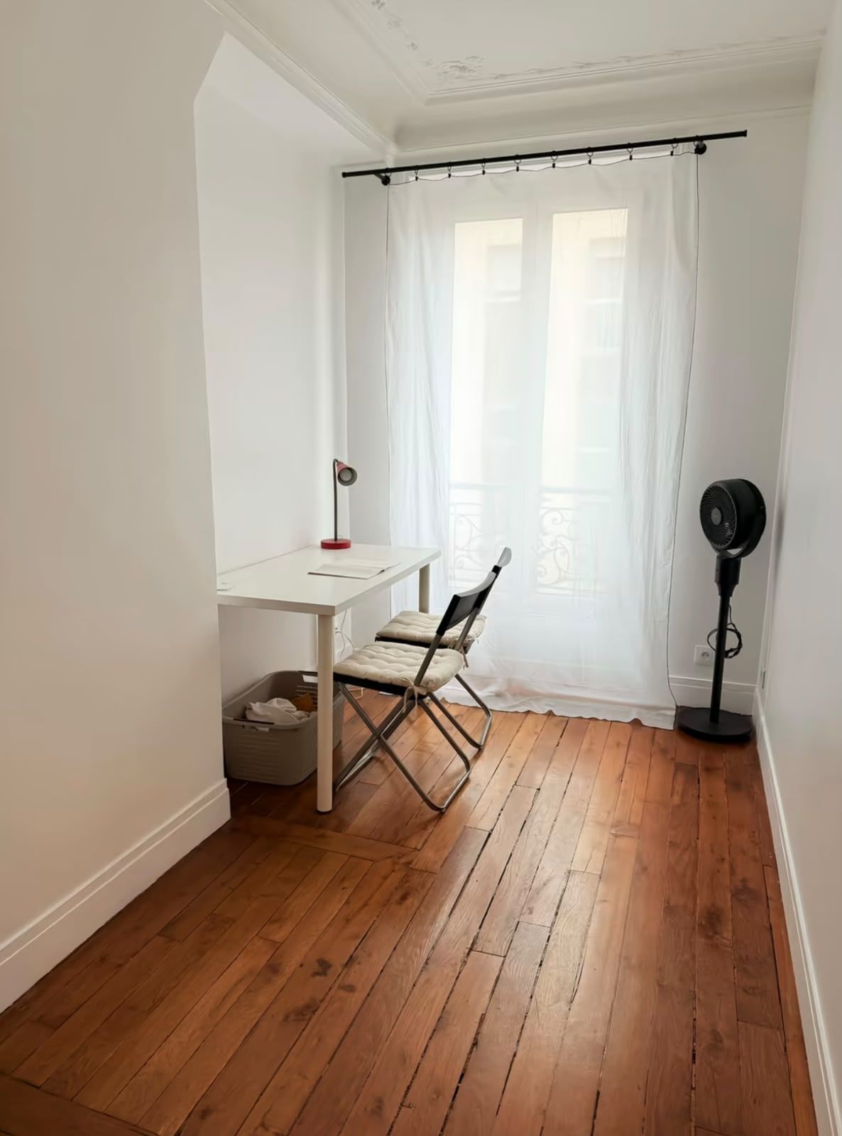 A room with a white table, two chairs, a floor fan, and a window with sheer curtains.