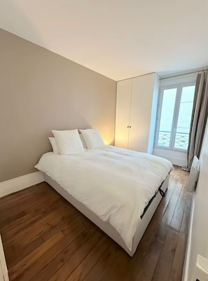 A bedroom with a bed, wooden flooring, a wardrobe, and a window with curtains. - Vrbo Property (Paris)