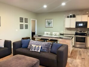 A modern living room with a sofa, a glass-top dining table, and a kitchen area with white cabinets and a stainless steel oven.