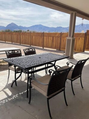 A covered patio with a metal table and chairs, a wooden fence, and mountains in the background.