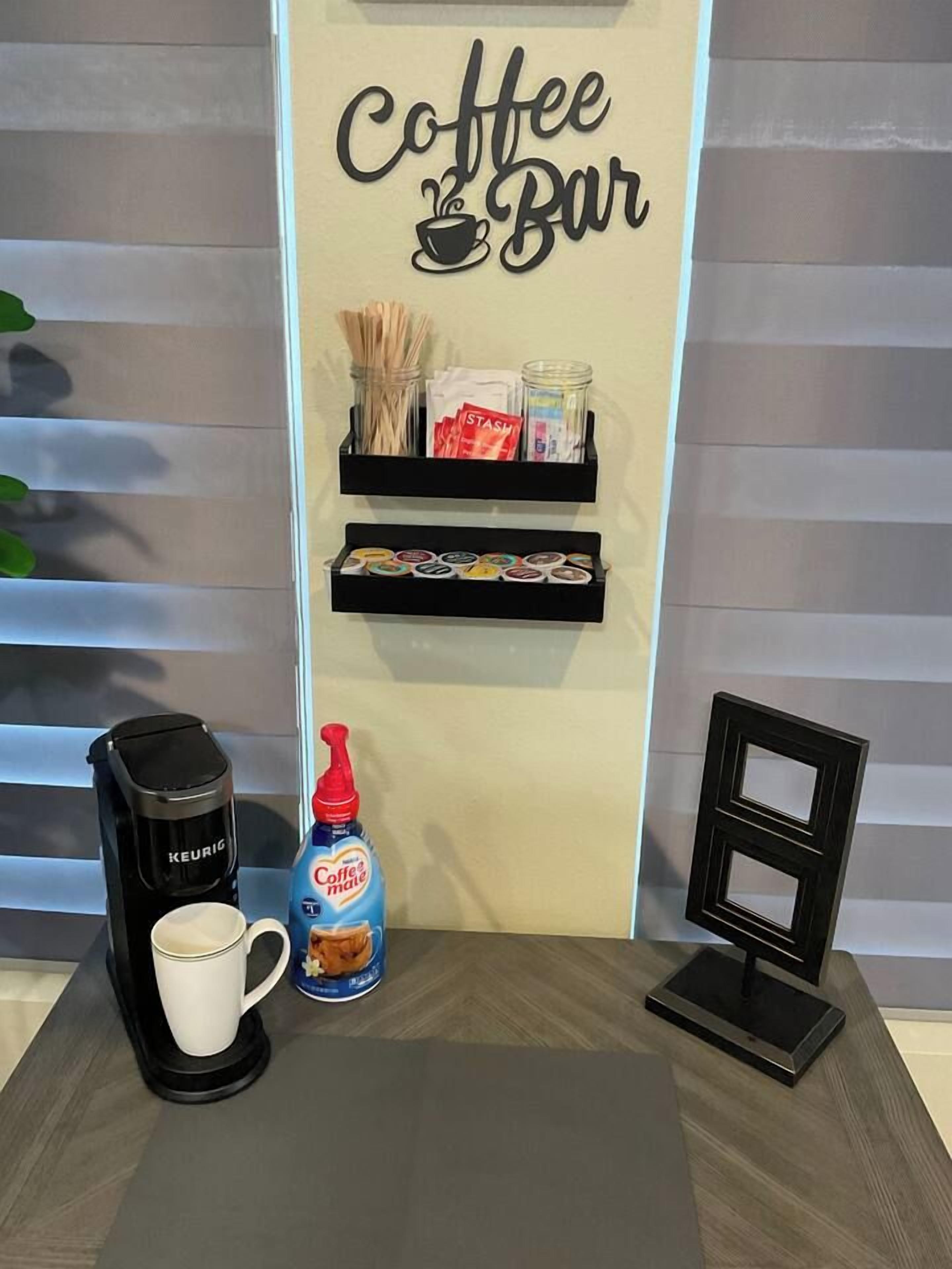 A coffee bar setup with a Keurig coffee maker, a mug, a bottle of coffee creamer, and a display shelf with coffee pods and snacks.