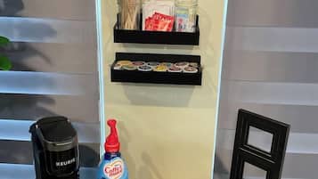 A coffee bar setup with a Keurig coffee maker, a mug, a bottle of coffee creamer, and a display shelf with coffee pods and snacks.