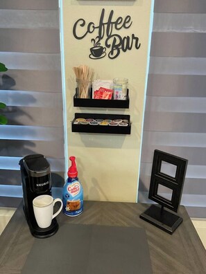 A coffee bar setup with a Keurig coffee maker, a mug, a bottle of coffee creamer, and a display shelf with coffee pods and snacks.
