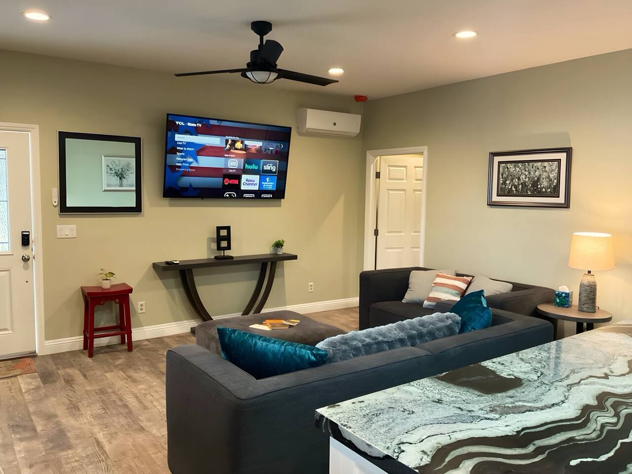 A living room with a flat-screen TV mounted on the wall, a sofa with cushions, a coffee table, and a small side table with a lamp.