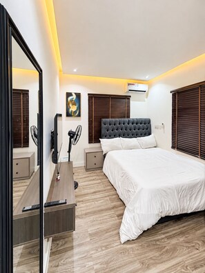 A modern bedroom with a large bed, a bedside table, a desk with a chair, a mirror, and a wall-mounted air conditioner. - 3 BEDROOM APARTMENT IN LIFECAMP GWARINPA ABUJA (abuja)