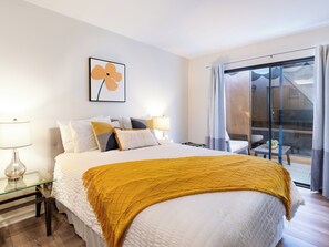 A hotel room with a bed, a glass table, a lamp, a wall art piece, and a view through a sliding glass door. - Modern Poolside 2 BR + Flex Space, Golf + Club Access (Palm Desert)