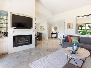 A modern living room with a fireplace, a flat-screen TV, a dining area, and a large window offering a view of the outdoors. - Modern Poolside 2 BR + Flex Space, Golf + Club Access (Palm Desert)