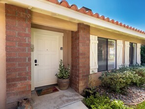 A residential building with a brick facade, a white door, and a covered porch area with potted plants and a doormat. - Modern Poolside 2 BR + Flex Space, Golf + Club Access (Palm Desert)