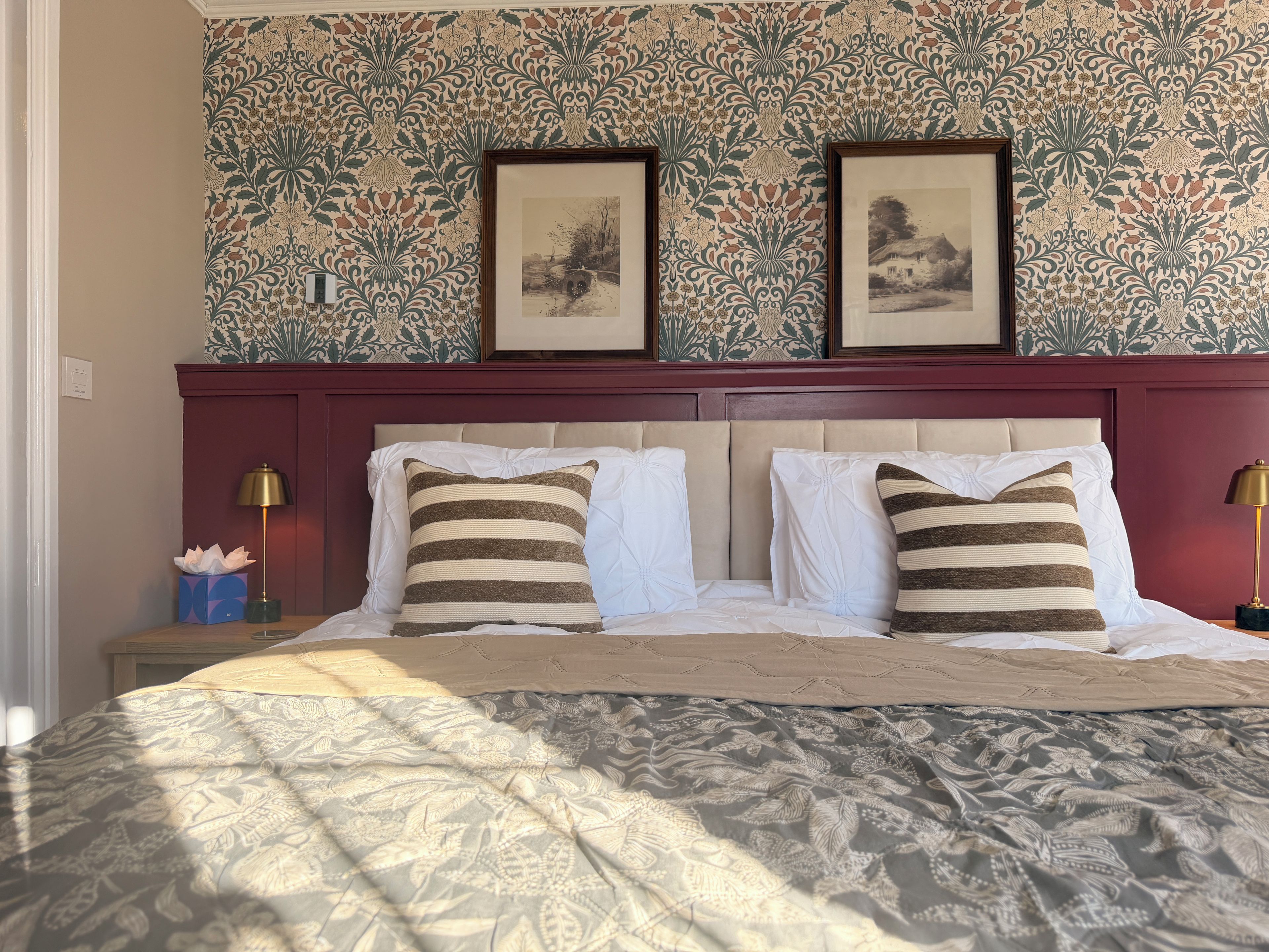 Una cama bien hecha con almohadas a rayas y una colcha estampada, una mesita de noche con lámpara, y una pared con papel tapiz floral y cuadros enmarcados.
