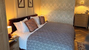 A well-appointed bedroom with a large bed, a nightstand with a lamp, a dresser, and a wall-mounted light fixture. - the Jenyns  (Denver)