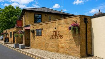 Un edificio de ladrillo con la señalización 'THE JENYNS', un letrero de bienvenida y macetas con flores en la pared.