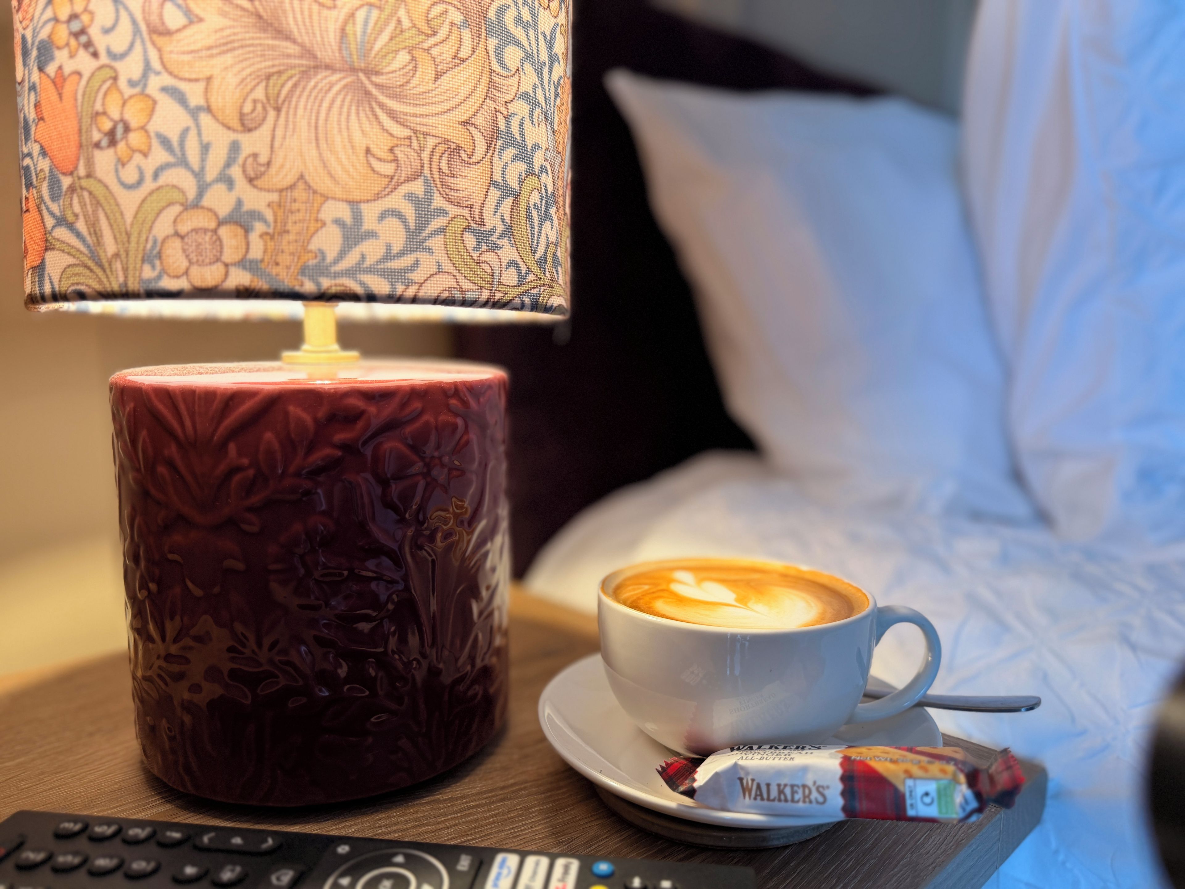 A cup of coffee with latte art, a remote control, a Walker's chocolate bar, and a patterned lamp on a bedside table.