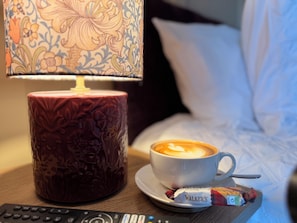 Una taza de café con arte de latte, un control remoto, una barra de chocolate Walker y una lámpara con un diseño sobre una mesita de noche.