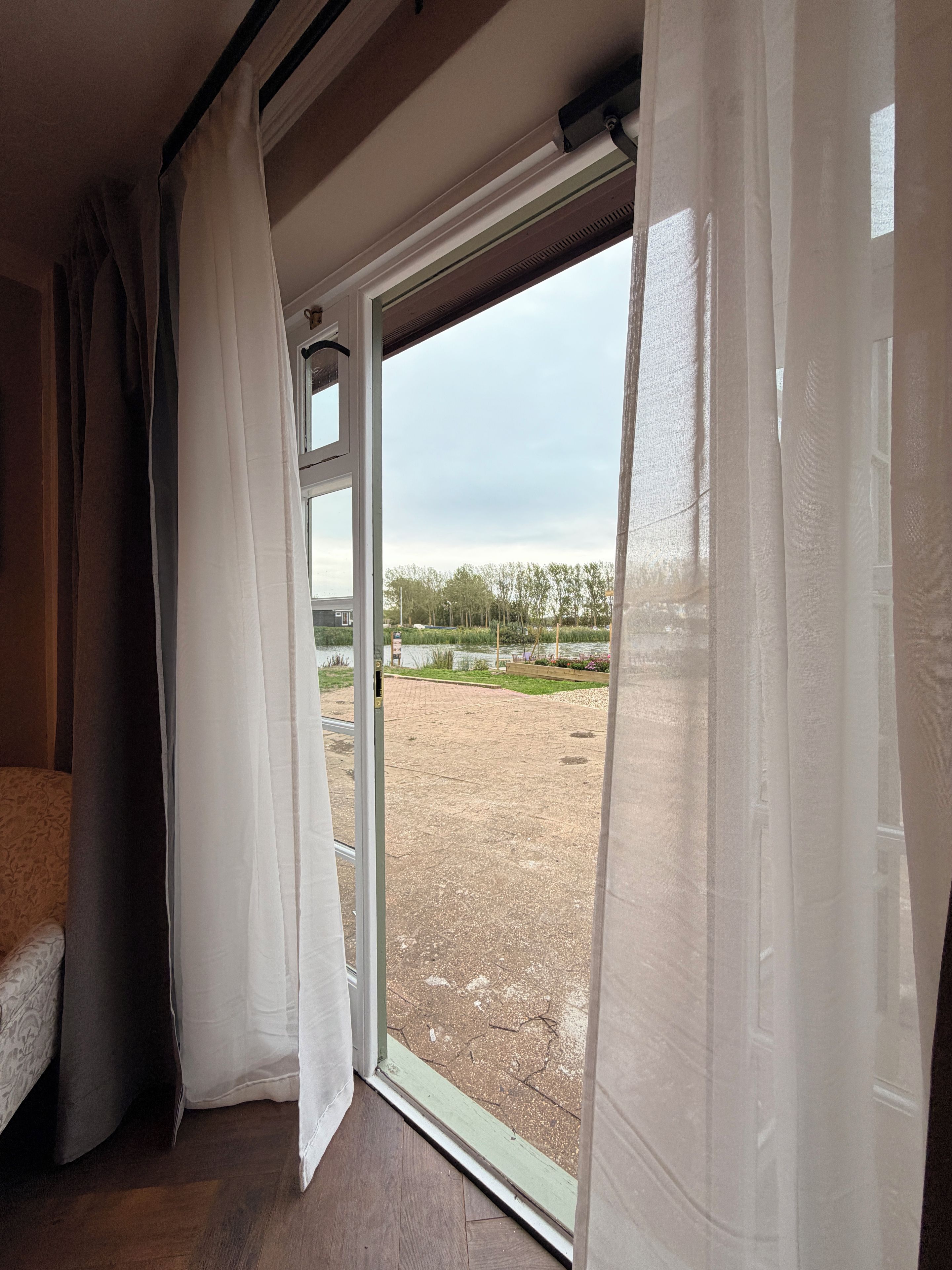 A room with a bed, a window with curtains, and a view of an outdoor area with trees and a paved path.