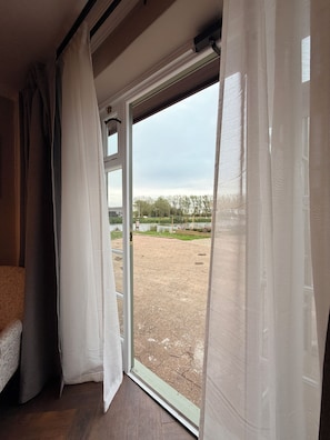 Una habitación con cama, ventana con cortinas y vista a un espacio exterior con árboles y un sendero pavimentado.