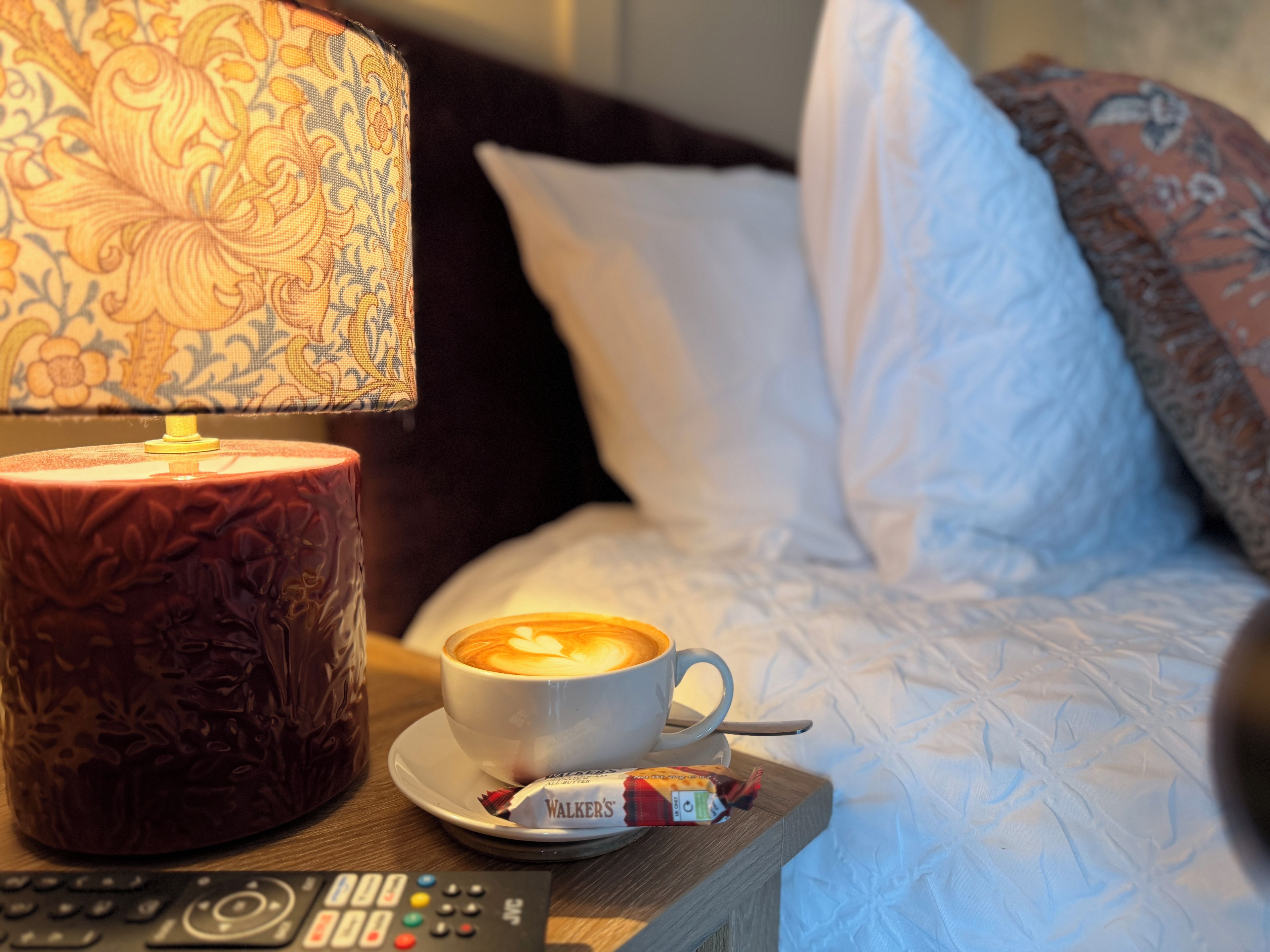 A bed with white bedding, a coffee cup with latte art, a remote control, and a packet of Walker's chocolate.