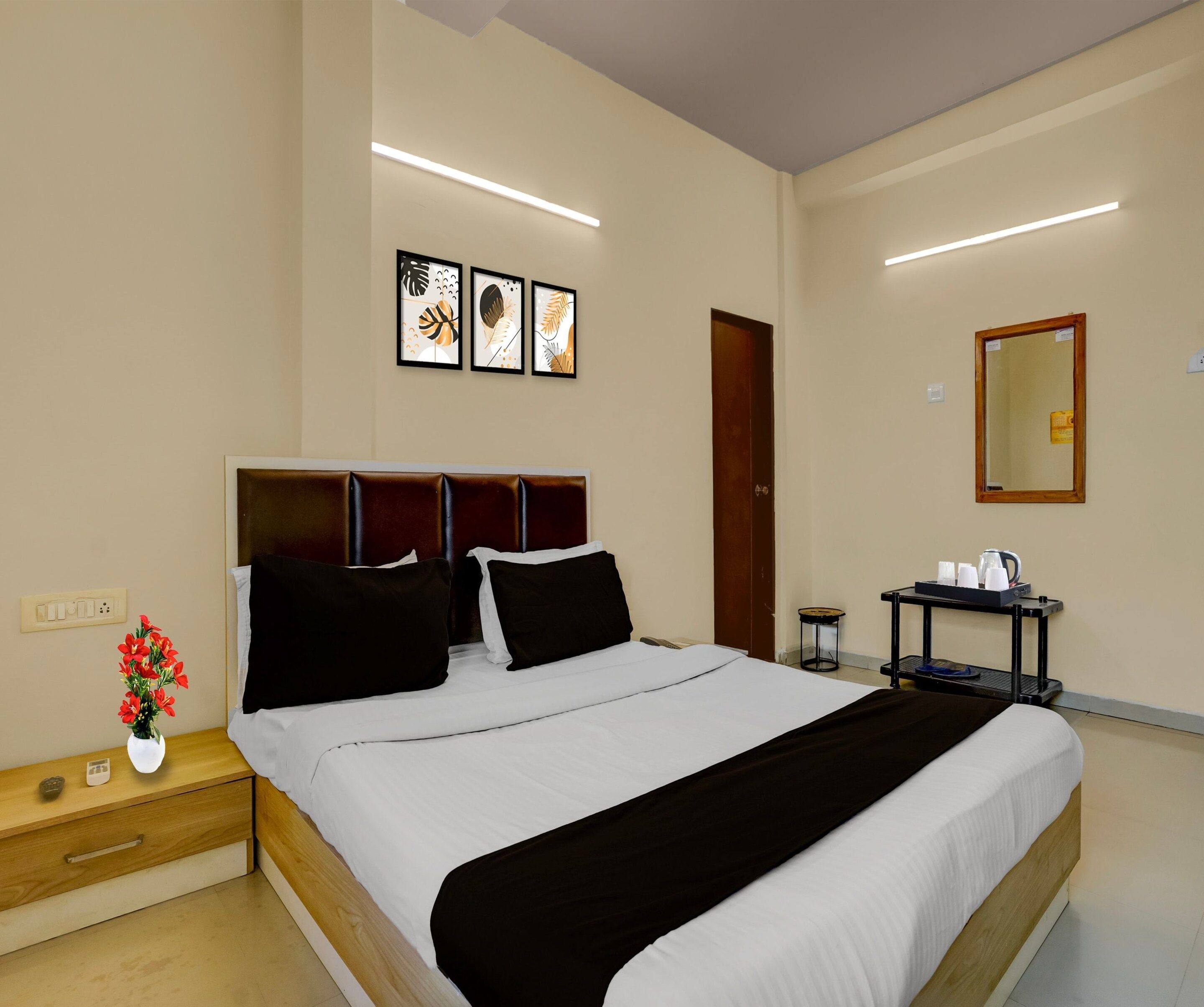 A hotel room with a bed, bedside table, a vase with flowers, a bedside lamp, a wall-mounted mirror, and a small table with cups and a kettle.