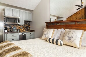 A bedroom with a bed, pillows, a wooden headboard, a kitchen area with appliances, and a ceiling fan.