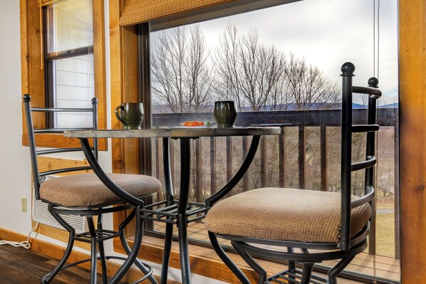 A balcony with a metal table and two chairs, overlooking a landscape with trees and a distant mountain range.