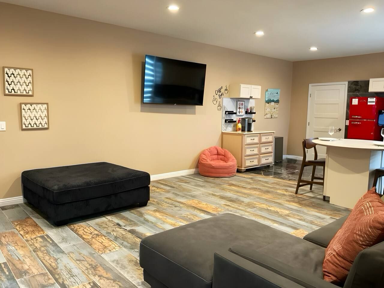 A living room with a flat-screen TV, a sofa, a bean bag, and a kitchen area with a refrigerator and cabinets.